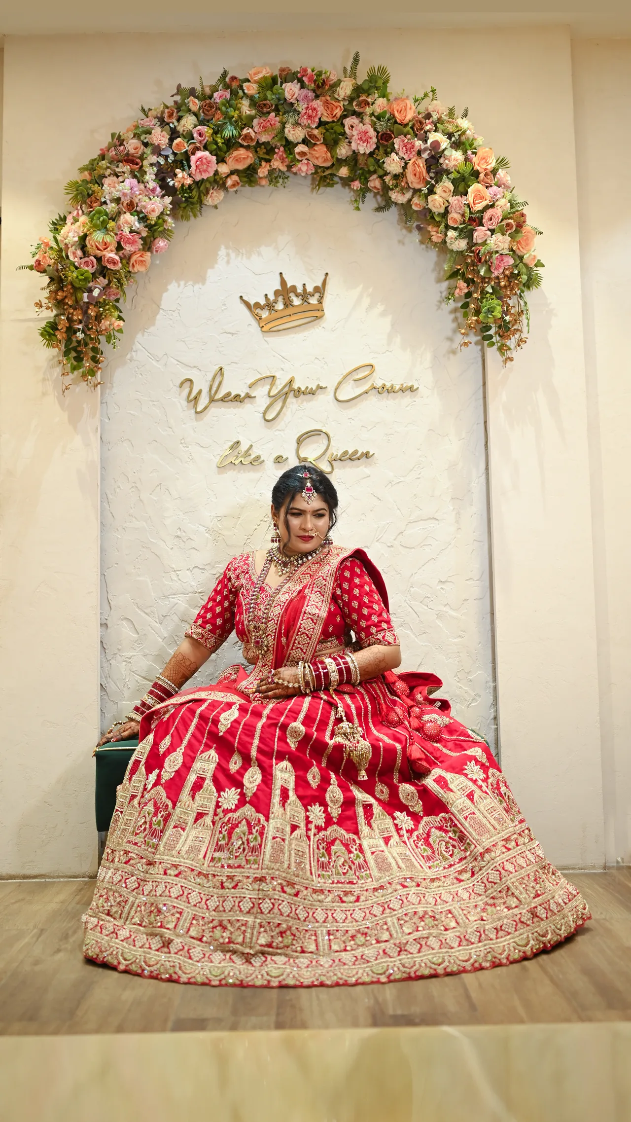Bride seated in a red lehenga beneath a floral arch