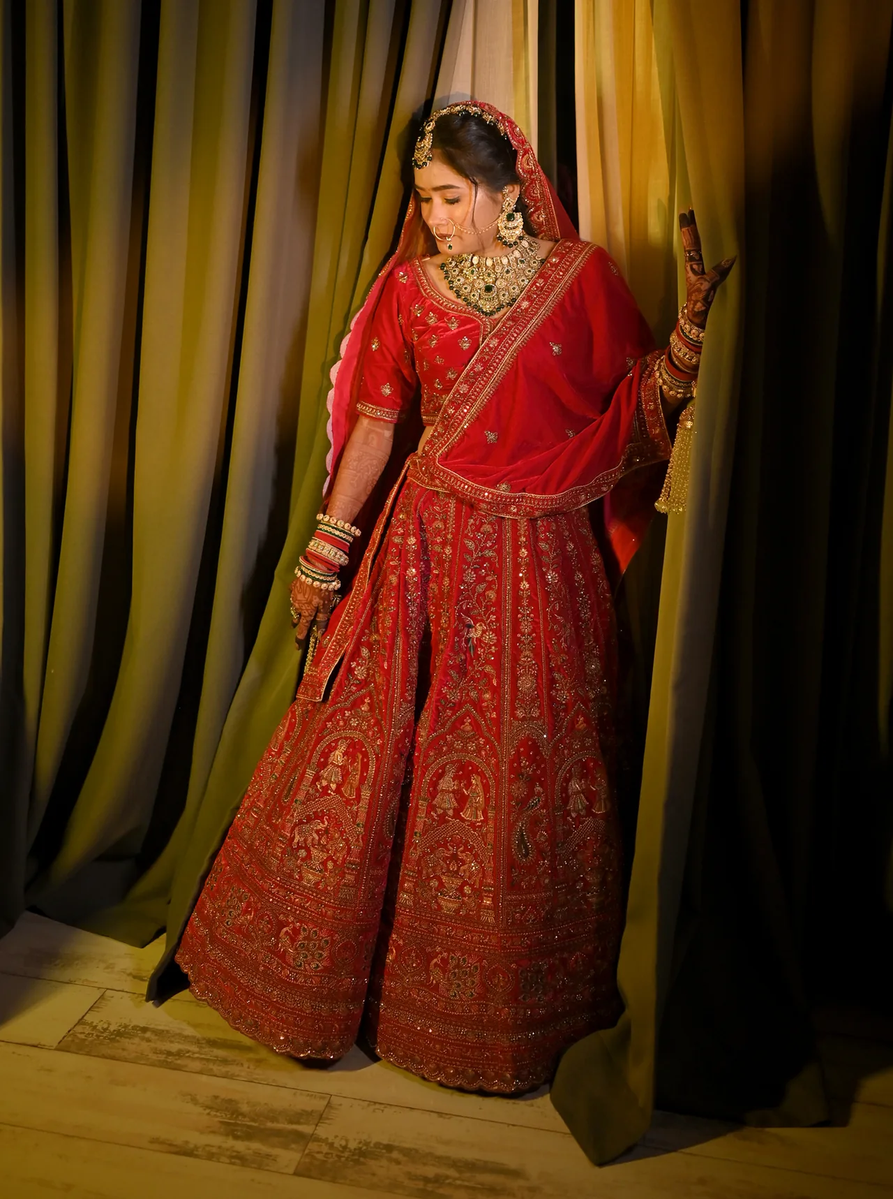 Bride in a red lehenga standing between olive curtains