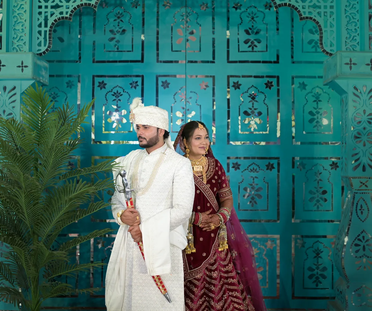 Groom in an ivory sherwani and bride in maroon before an ornate teal door